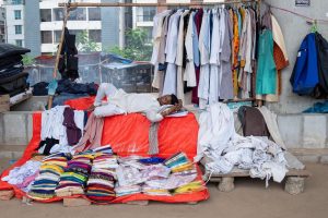 A street vendor naps on a market stall surrounded by colorful clothing in an urban setting.